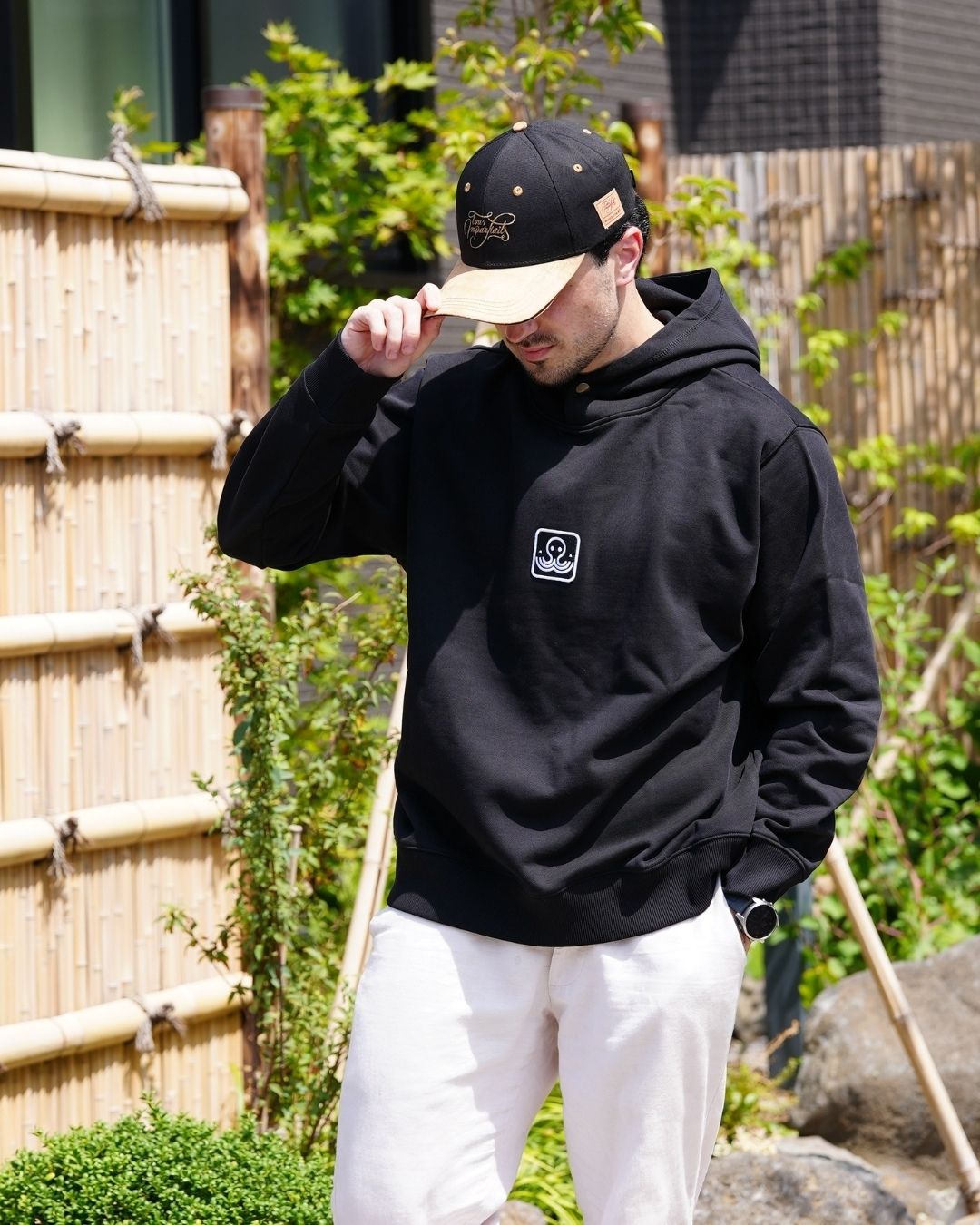 Modèle masculin portant le hoodie Basics noir en extérieur, en tenant sa casquette noire Tous Imparfaits