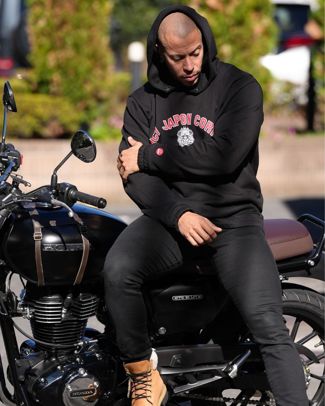 JC assis sur sa moto, portant le hoodie University noir, avec la capuche